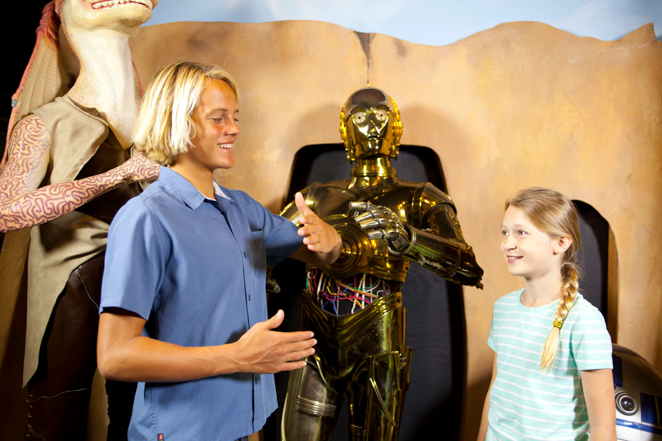 A boy and a girl stand next to a golden robot, smiling and gesturing as they interact.