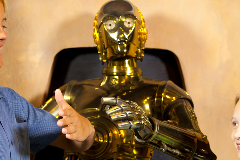 A boy and a girl stand next to a golden robot, smiling and gesturing as they interact.