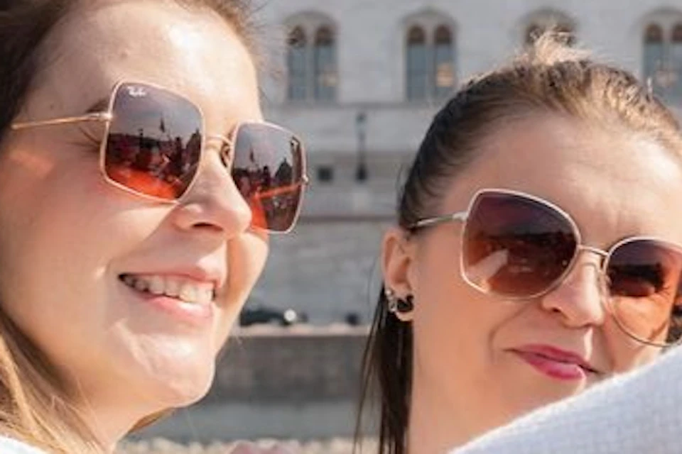 Two women take a selfie on a boat with the Hungarian Parliament Building in the background on a sunny day.