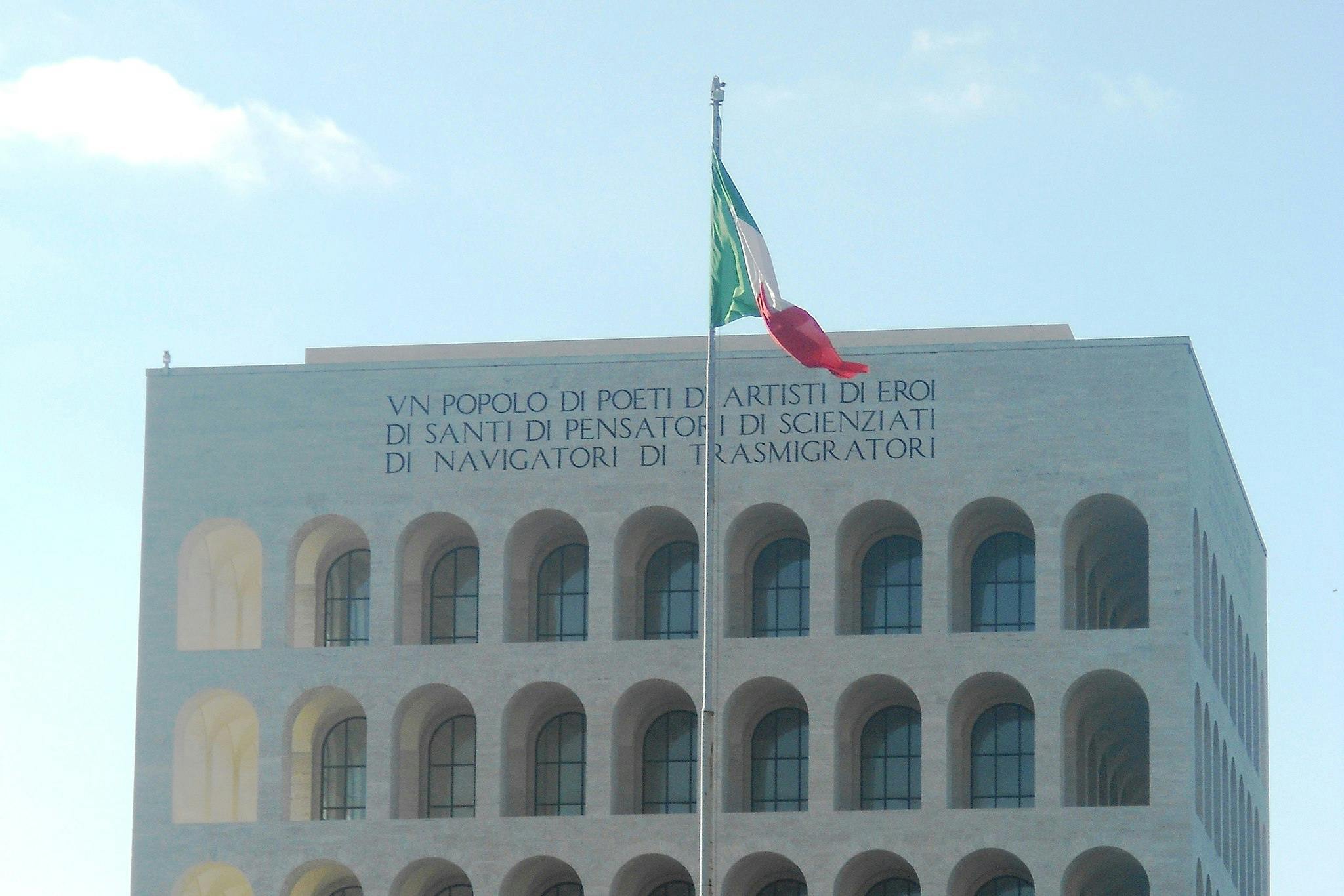 Italian rectangular building with repetitive arches, an Italian flag, and inscription "UN POPOLO DI POETI... TRASMIGRATORI" on top.