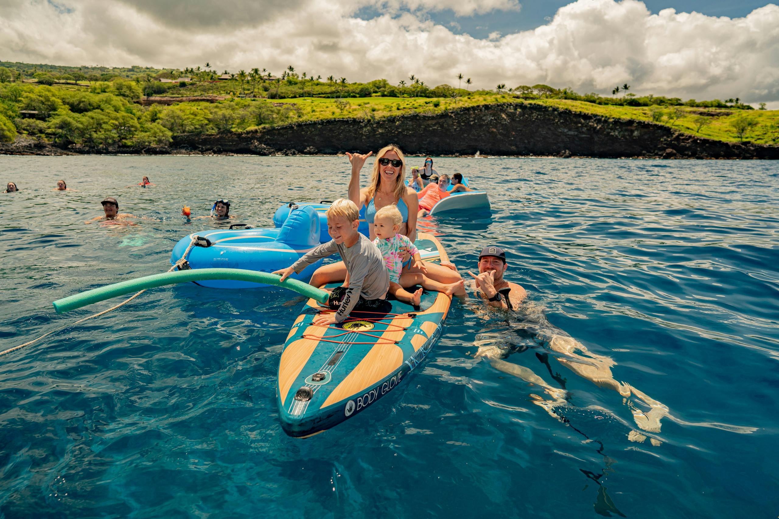 People enjoying a sunny day on a lake, with adults and children on paddleboards and inflatables near the shore.