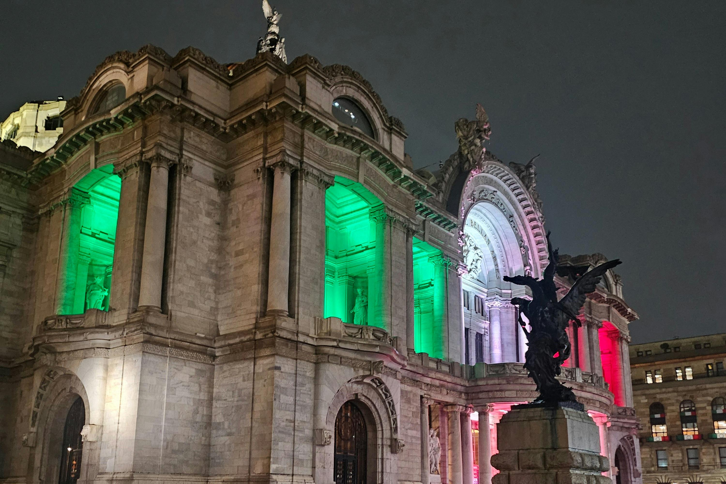Een historisch gebouw 's nachts verlicht met groene, witte en rode lichten, met een standbeeld op de voorgrond.