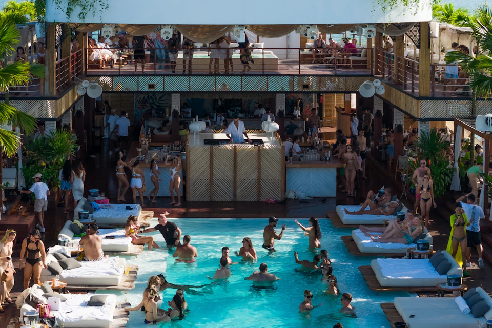 People enjoying a pool party with music, dancing, and lounging on white daybeds, featuring an upper and lower deck area.