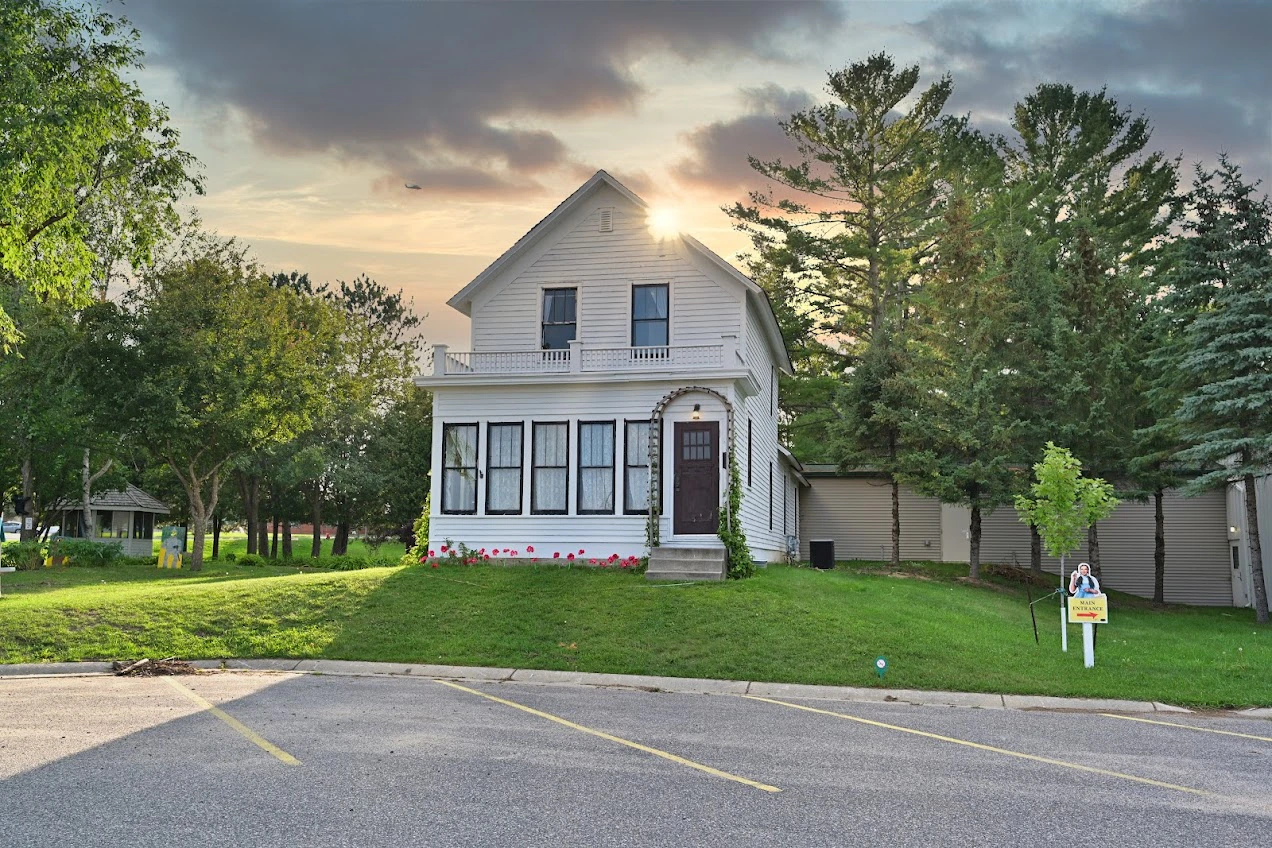 Two-story white house with a front porch, surrounded by green trees and a lawn, with a parking lot in the foreground.