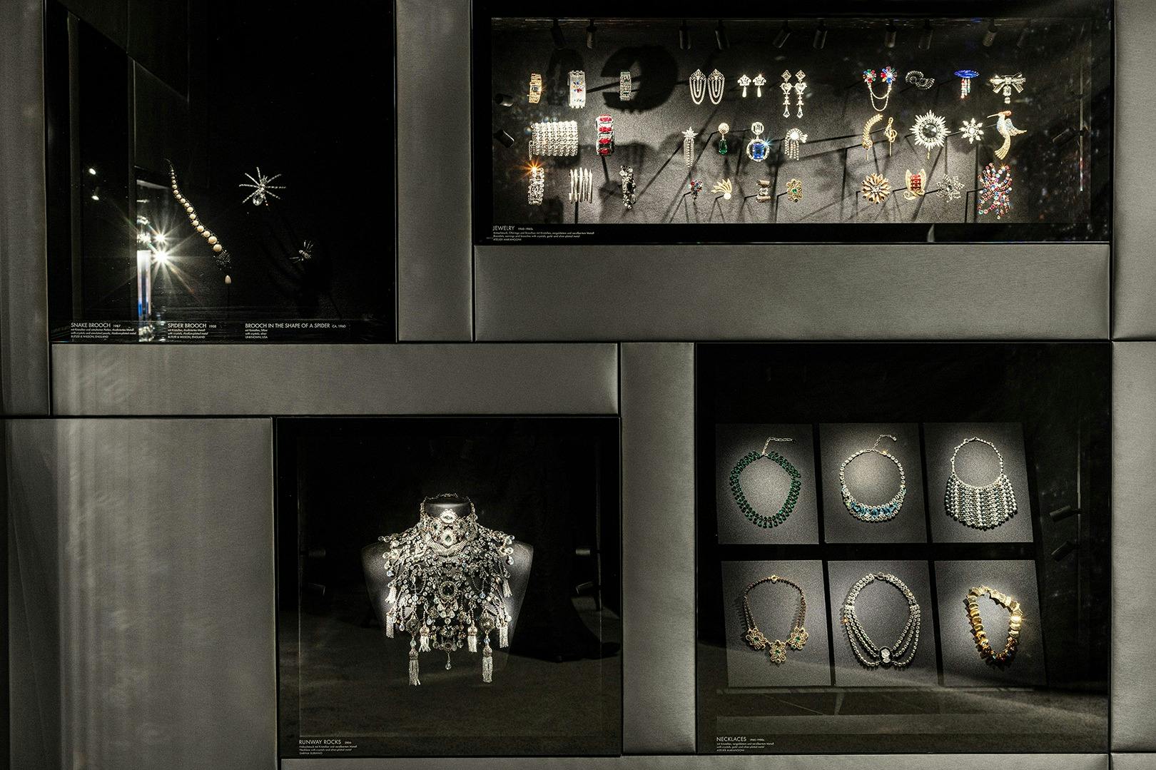 ガラスのディスプレイケースには、ブローチ、イヤリング、ブレスレット、ネックレスなど、さまざまなジュエリーが暗い背景にセットされています。