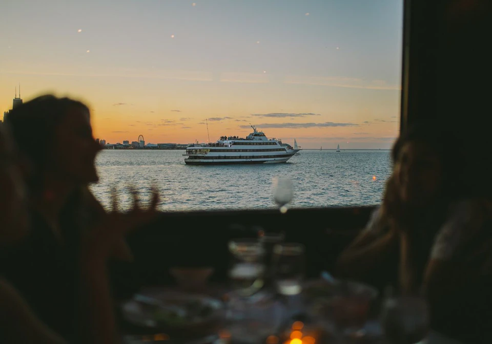 People dining indoors with blurred silhouettes in foreground; large boat on water, city skyline and sunset in background.