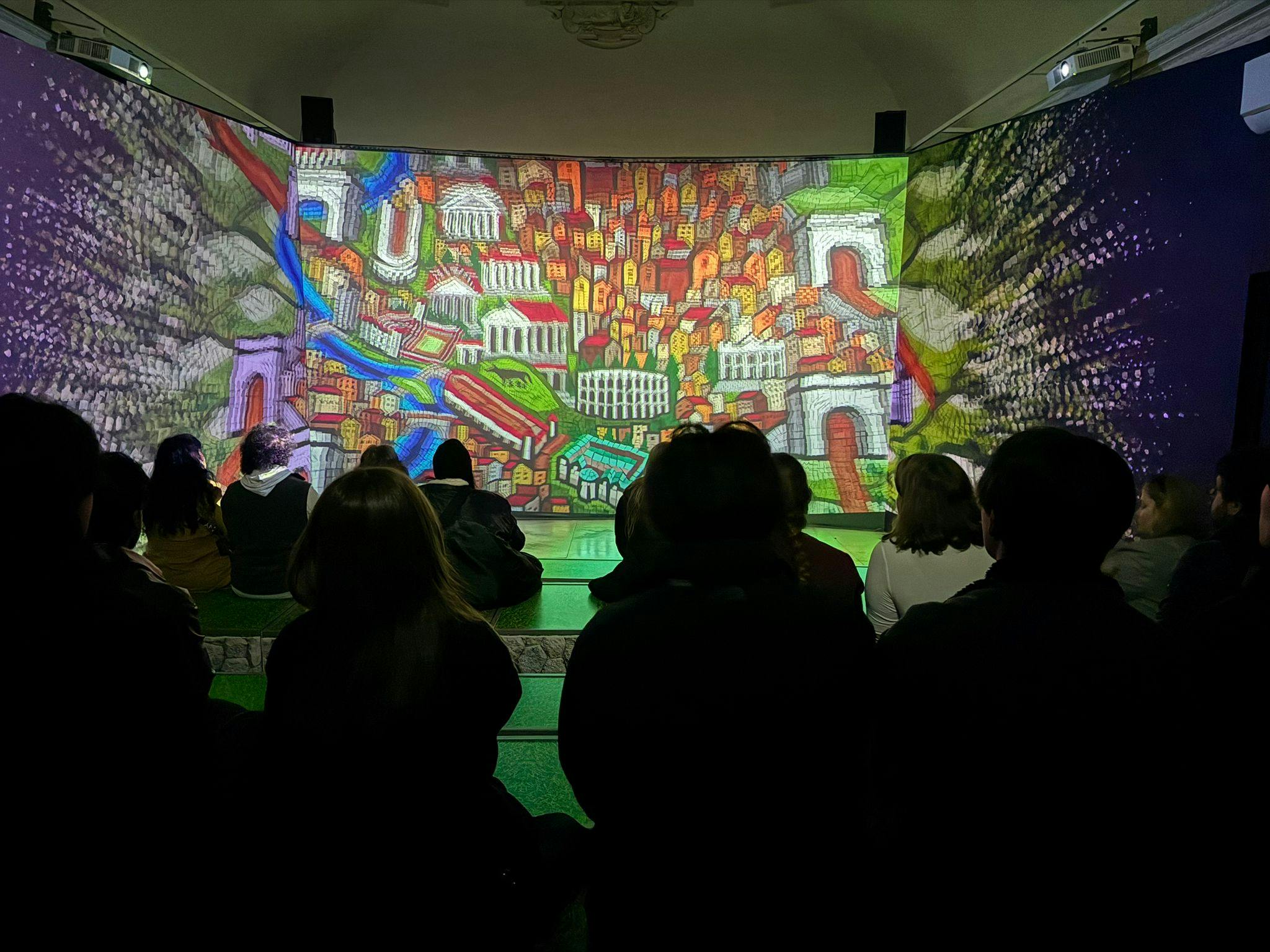 Persone sedute all'interno che guardano una vibrante e colorata proiezione di un paesaggio urbano su grandi schermi circostanti.