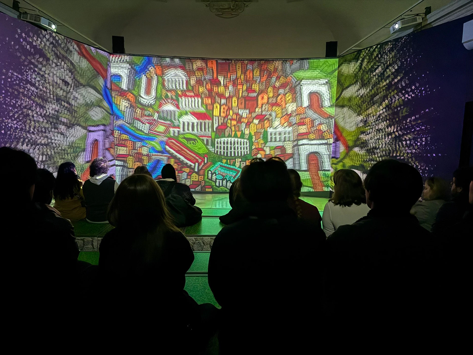 People seated indoors watching a vibrant and colorful projection of a cityscape on large surrounding screens.