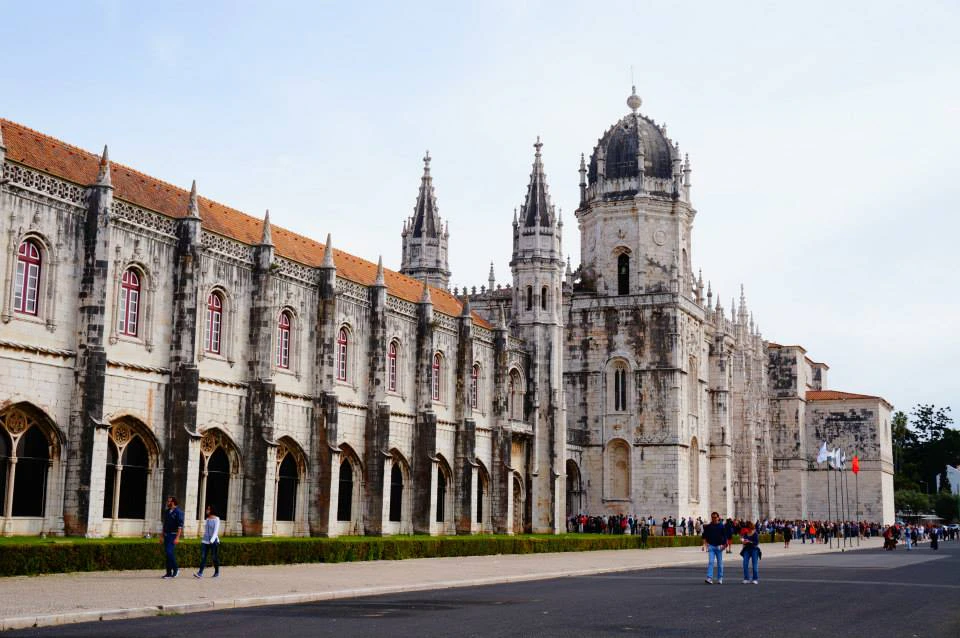 Historical stone building with pointed arches, ornate towers, and a domed structure. People walk on a paved path nearby.