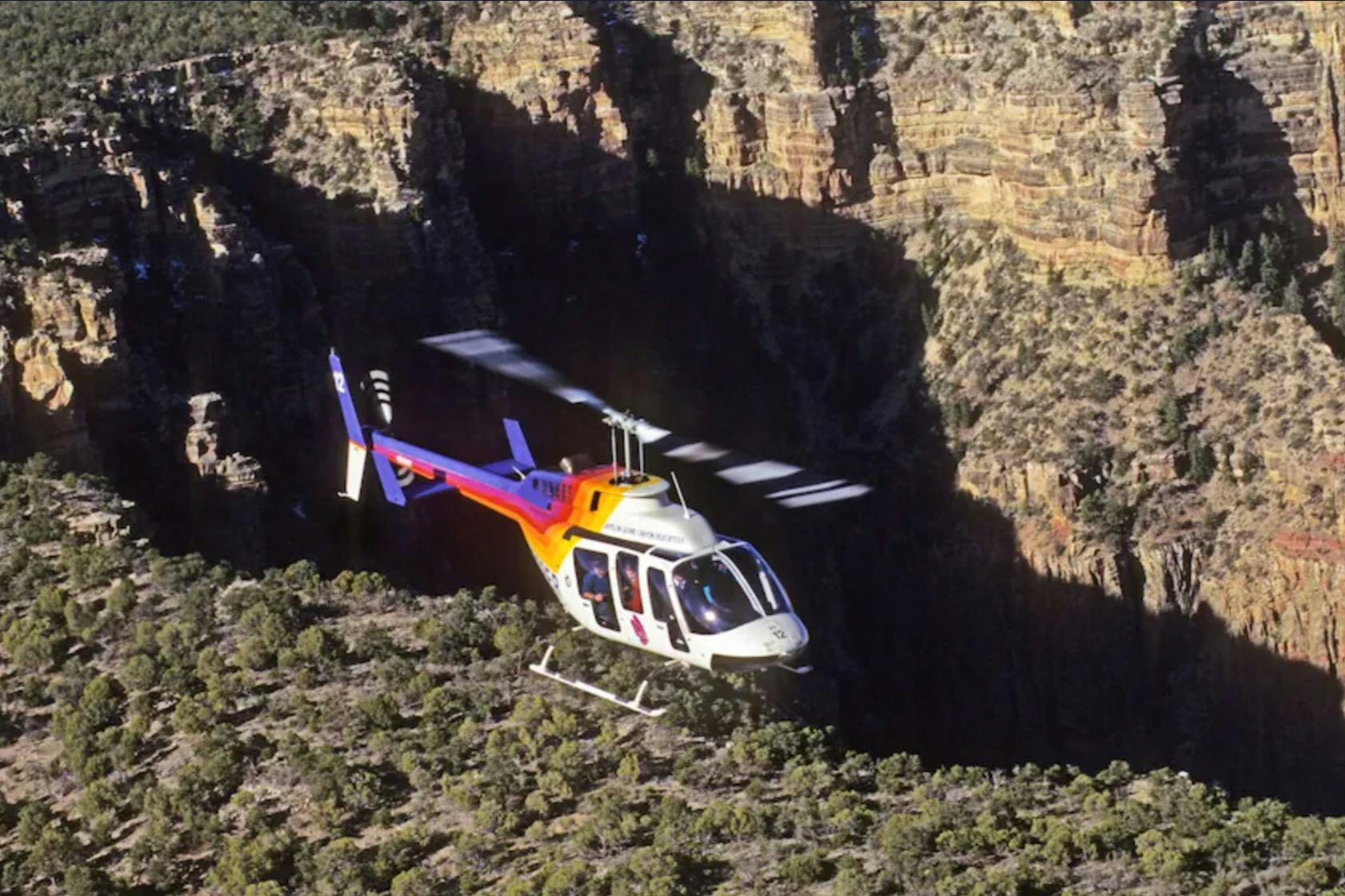 A helicopter with a white and orange design flying over a canyon surrounded by rocky cliffs and greenery.