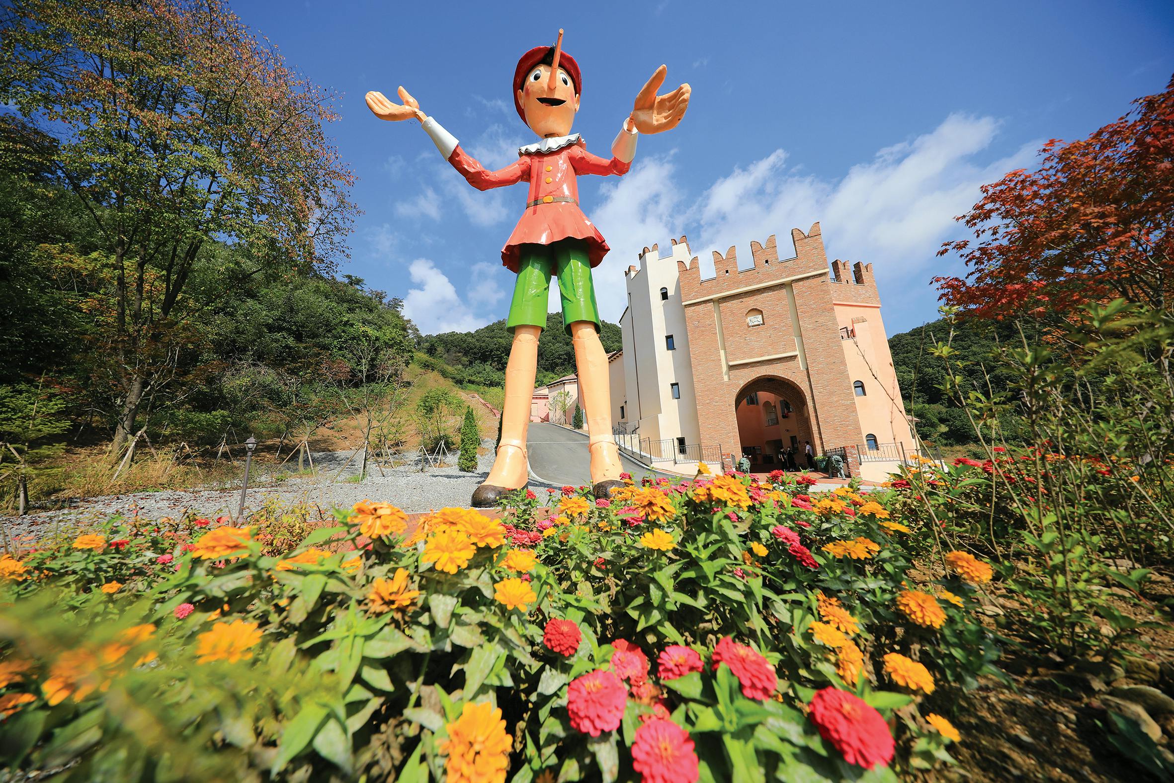 A large Pinocchio statue stands among colorful flowers in front of a castle-like building under a clear blue sky.