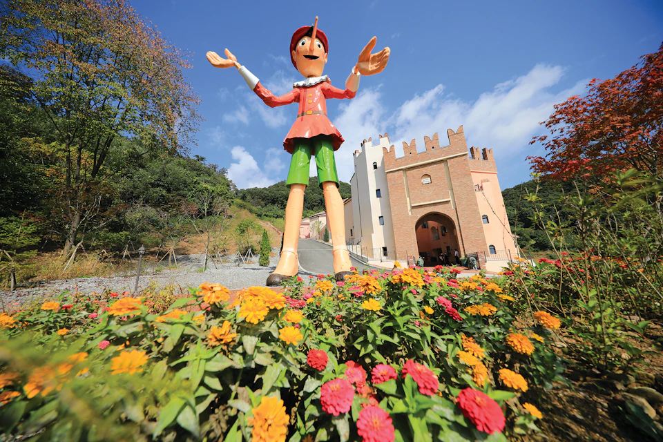A large Pinocchio statue stands among colorful flowers in front of a castle-like building under a clear blue sky.