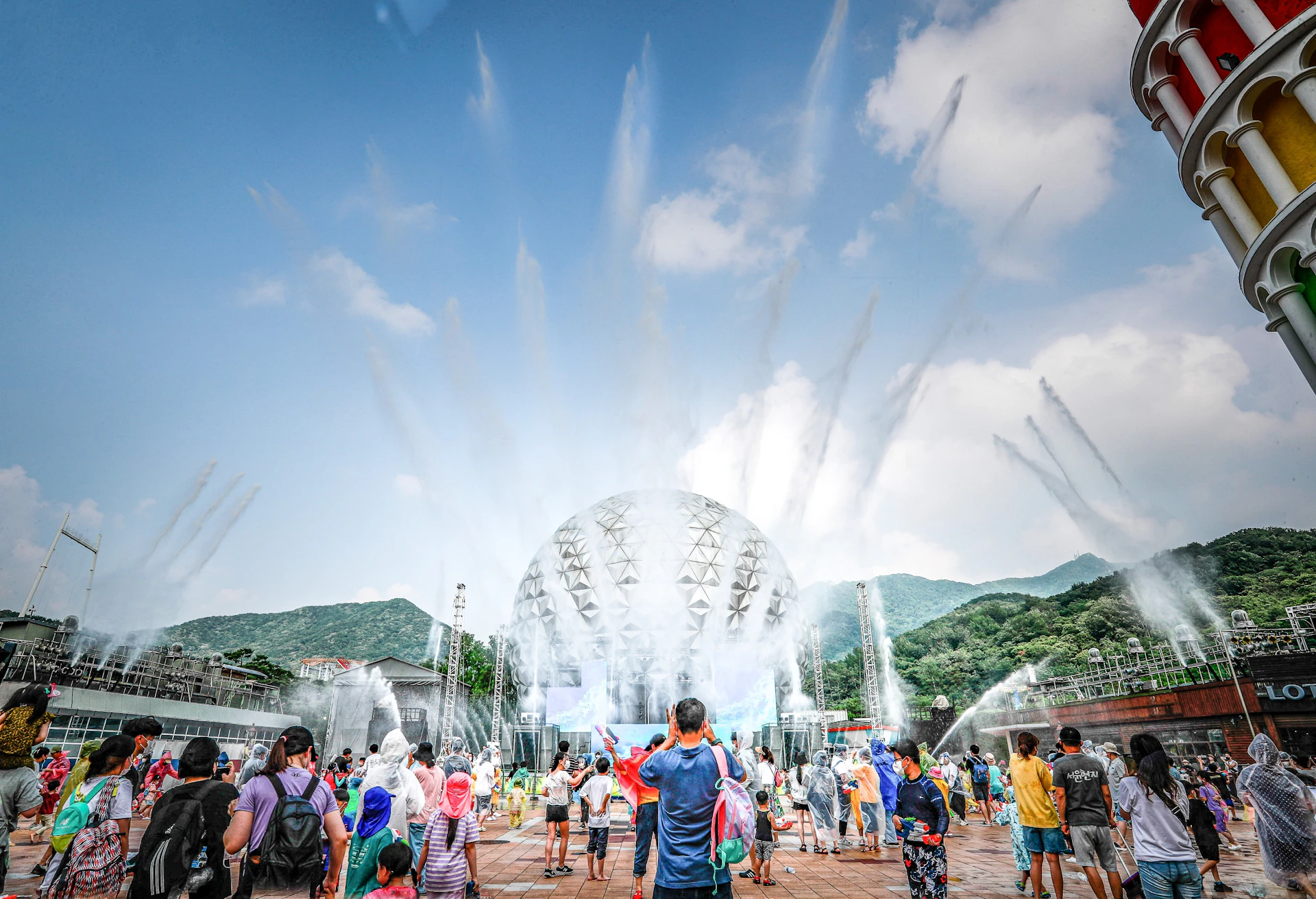 A crowd watches a water fountain show with jets in front of a spherical building surrounded by mountains under a blue sky.