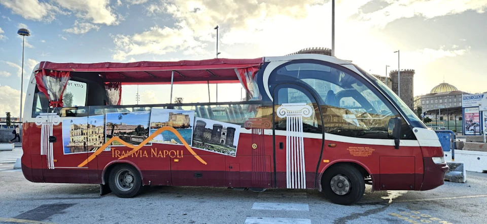 A red tourist bus with images of Naples landmarks, including castles and columns, parked on a sunny day.