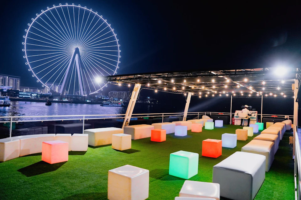 Night scene with colorful cube seats on a rooftop terrace, string lights above, and a large Ferris wheel in the background.
