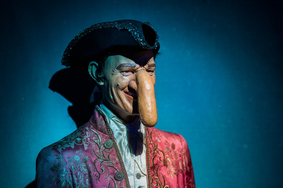 A figure in an ornate costume with a tricorne hat and exaggerated facial features, lit dramatically against a dark background.