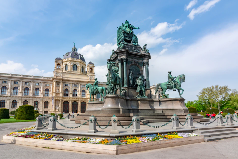 A grand statue with multiple equestrian figures in a plaza, in front of a historic domed building and vibrant flowerbeds.