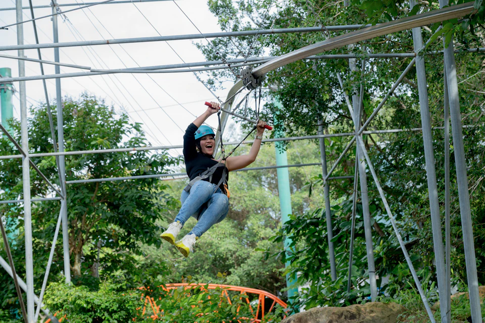 A person wearing safety gear and smiling while riding a zipline through a lush, green outdoor setting.
