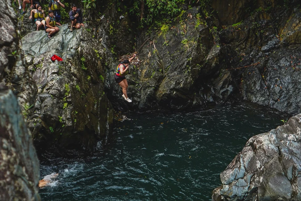 A person swinging from a rope over a rocky creek with onlookers perched on the rocks nearby.