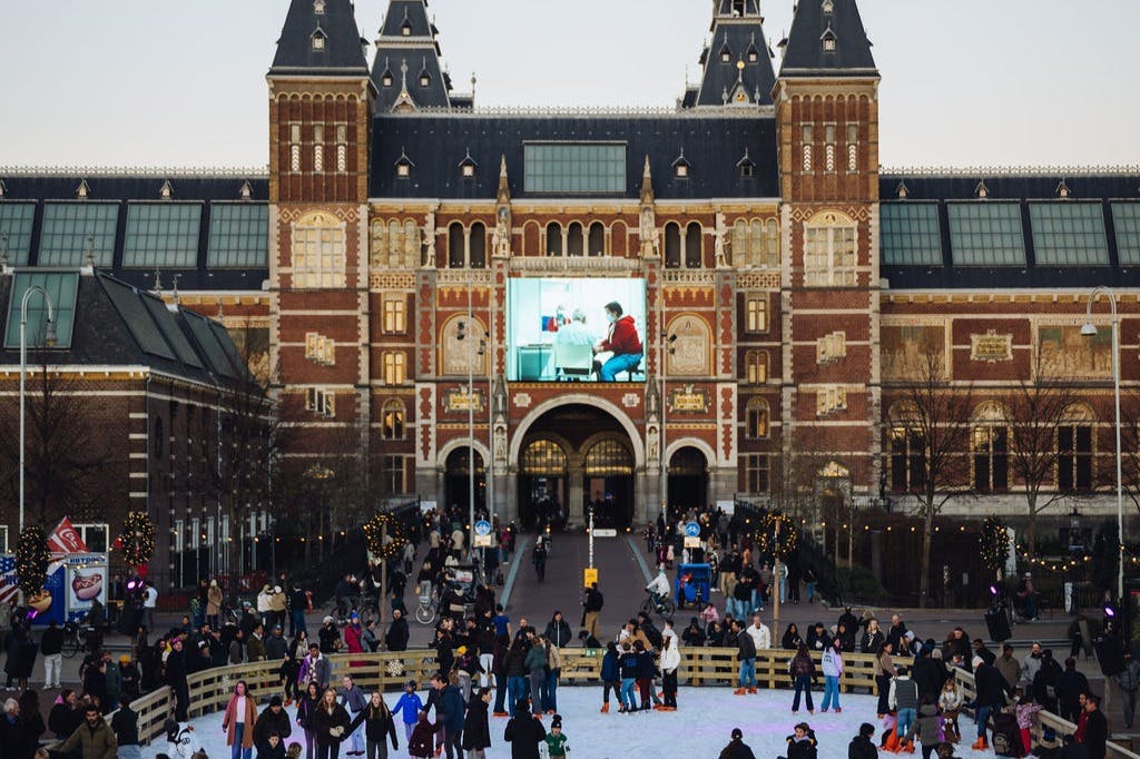 People ice skating in front of a large, ornate brick building with a screen displaying an image above the entrance.