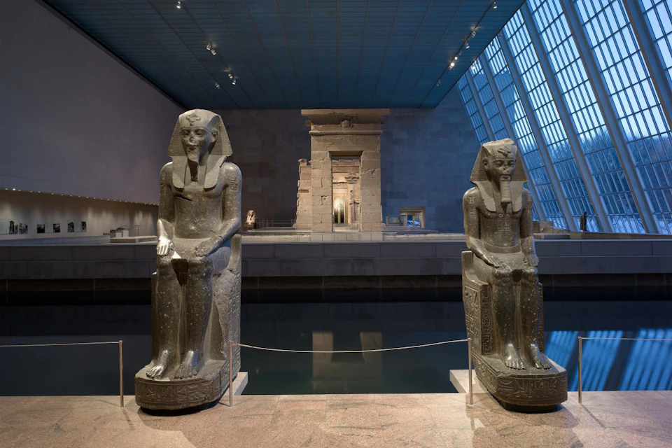 Two large ancient Egyptian statues sit facing forward in a museum, with a stone temple structure in the background.