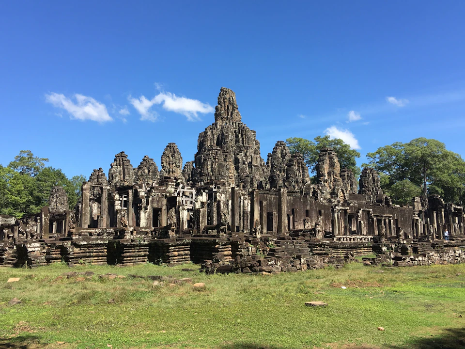 Ancient stone temple ruins with multiple towers under a clear blue sky, surrounded by grass and trees.