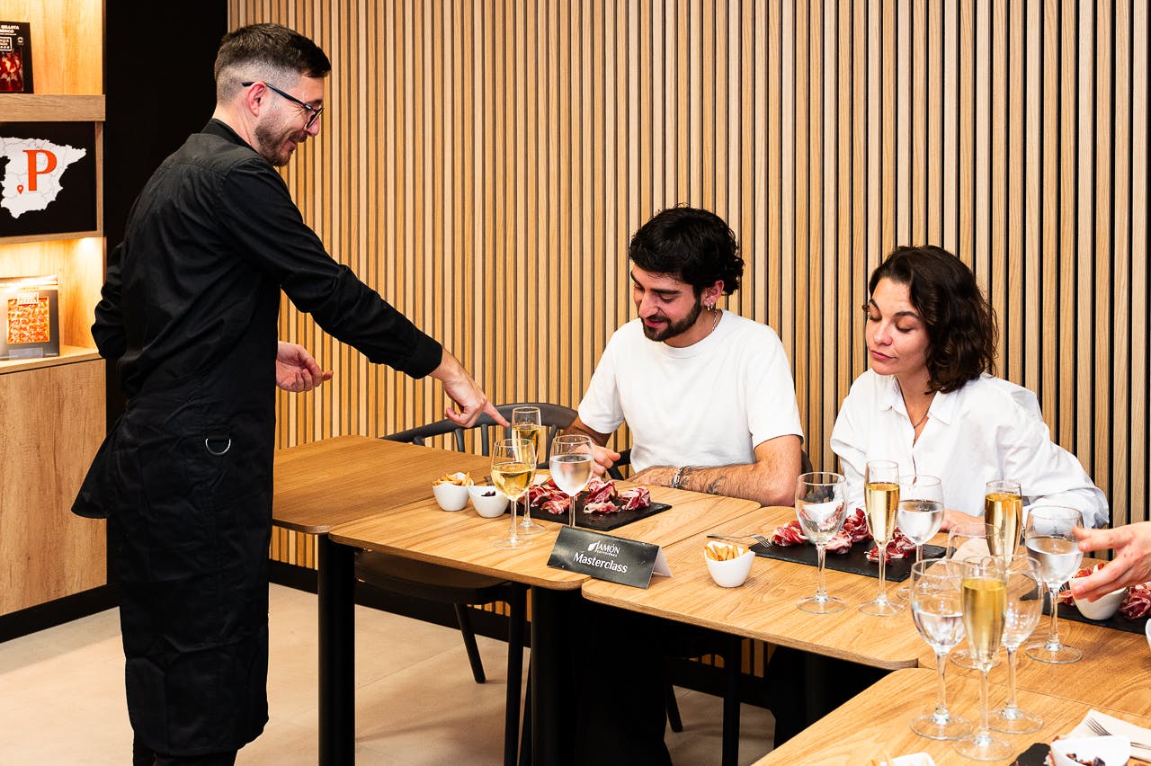 A man in a black apron interacts with two seated individuals at a table set with food and drinks in a modern interior.