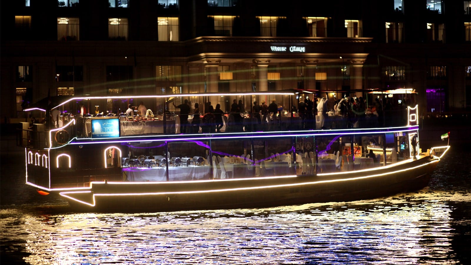 A boat illuminated with lights, filled with people, is docked near a brightly lit building with "Venice Plaza" signage visible.