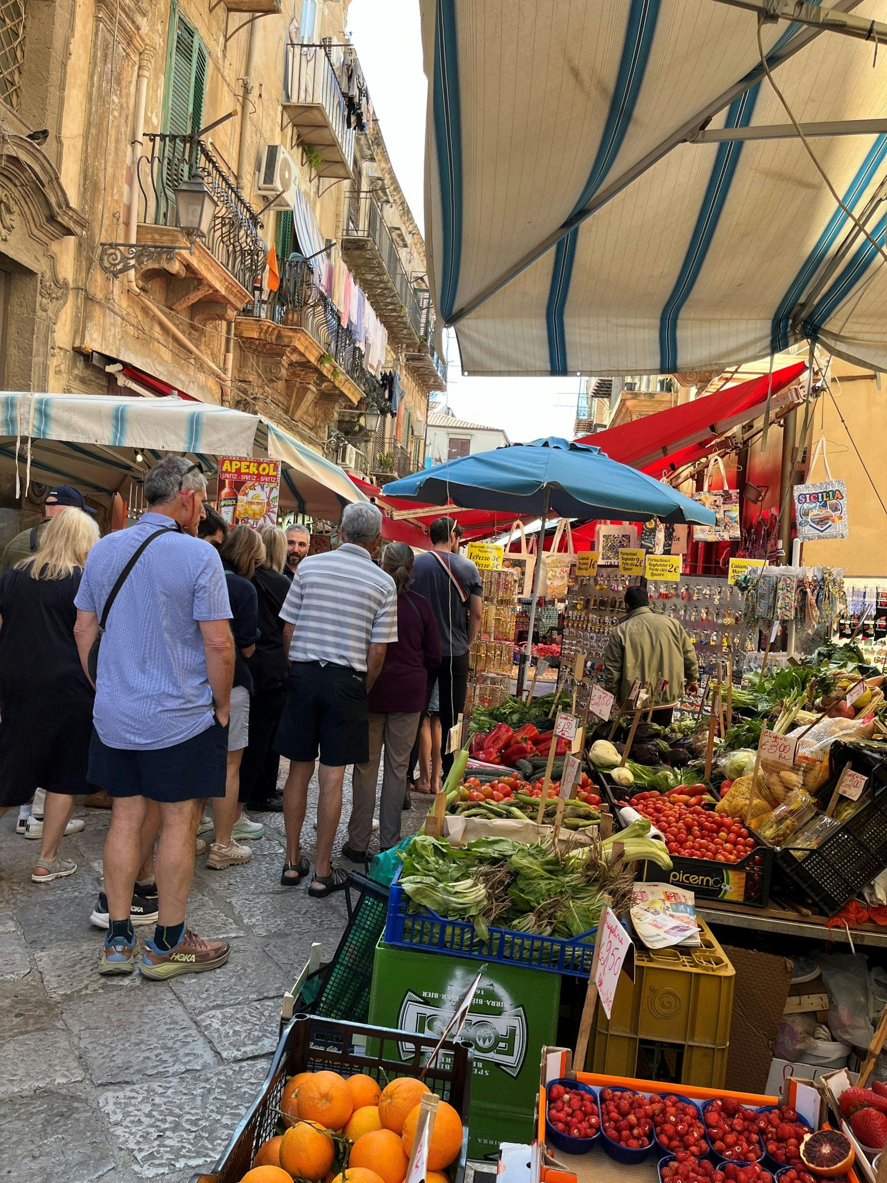 A bustling outdoor market with people browsing stalls filled with fresh produce under colorful umbrellas and awnings.