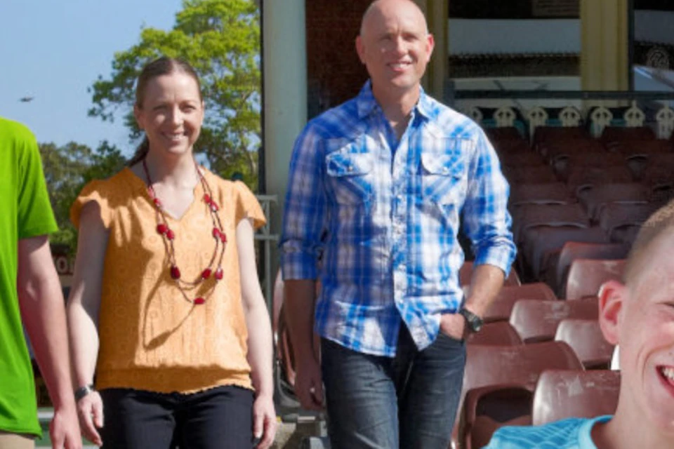 A group of four people outdoors, with three walking and one running in the foreground, near a building and seating area.
