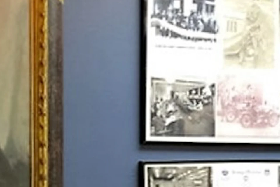 A man stands next to a glass display case with historical artifacts, in front of a large portrait and framed historical photographs on the wall.