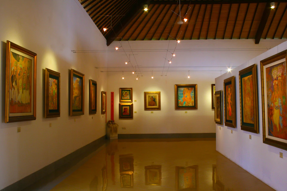 A small art gallery with framed paintings on white walls, wooden ceiling, tile floor, and ceiling lights illuminating the artworks.