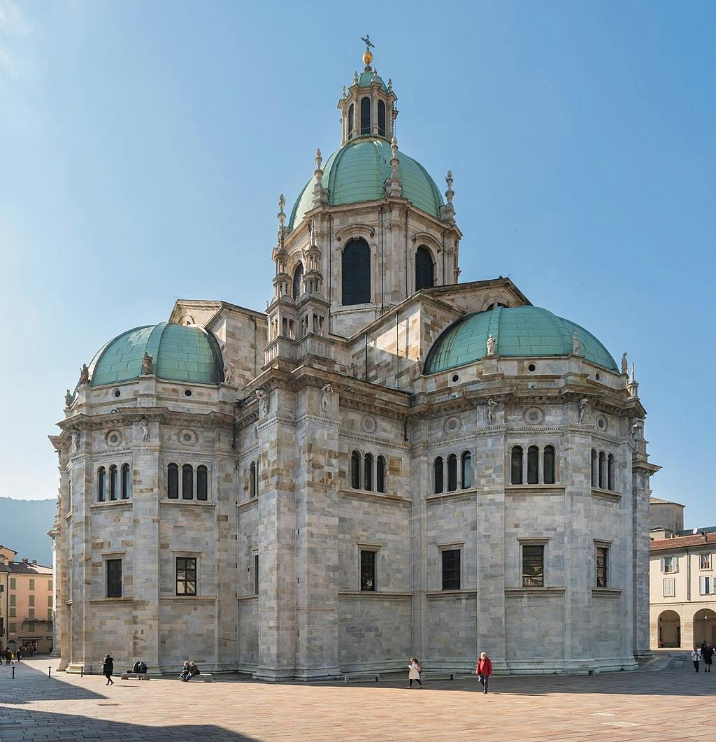 Un gran edifici històric de pedra amb múltiples cúpules, gent visible caminant i asseguda en una plaça oberta sota un cel blau clar.