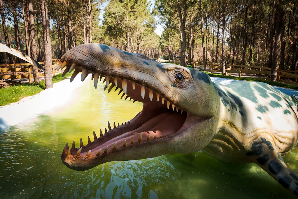 A life-sized dinosaur model with an open mouth and sharp teeth, in a green, water-filled area surrounded by trees.