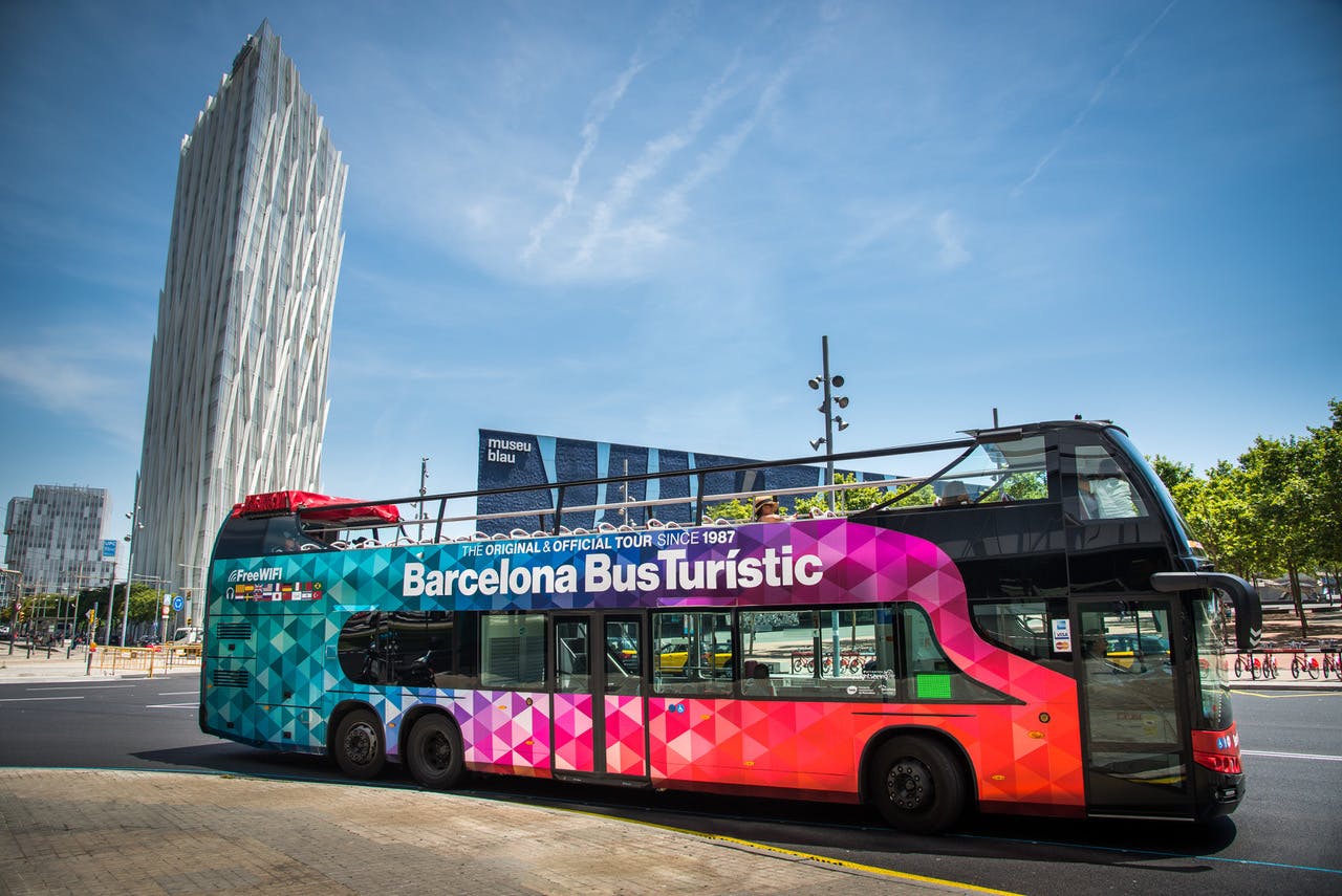 "Barcelona Bus Turistic" 이라는 이름의 다채로운 2층 투어 버스가 현대적인 건물 앞에 맑고 푸른 하늘 아래 주차되어 있습니다.