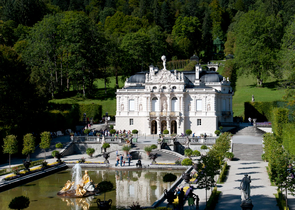 A grand palace with intricate architecture, a large fountain with statues in front, and people walking in a landscaped garden.