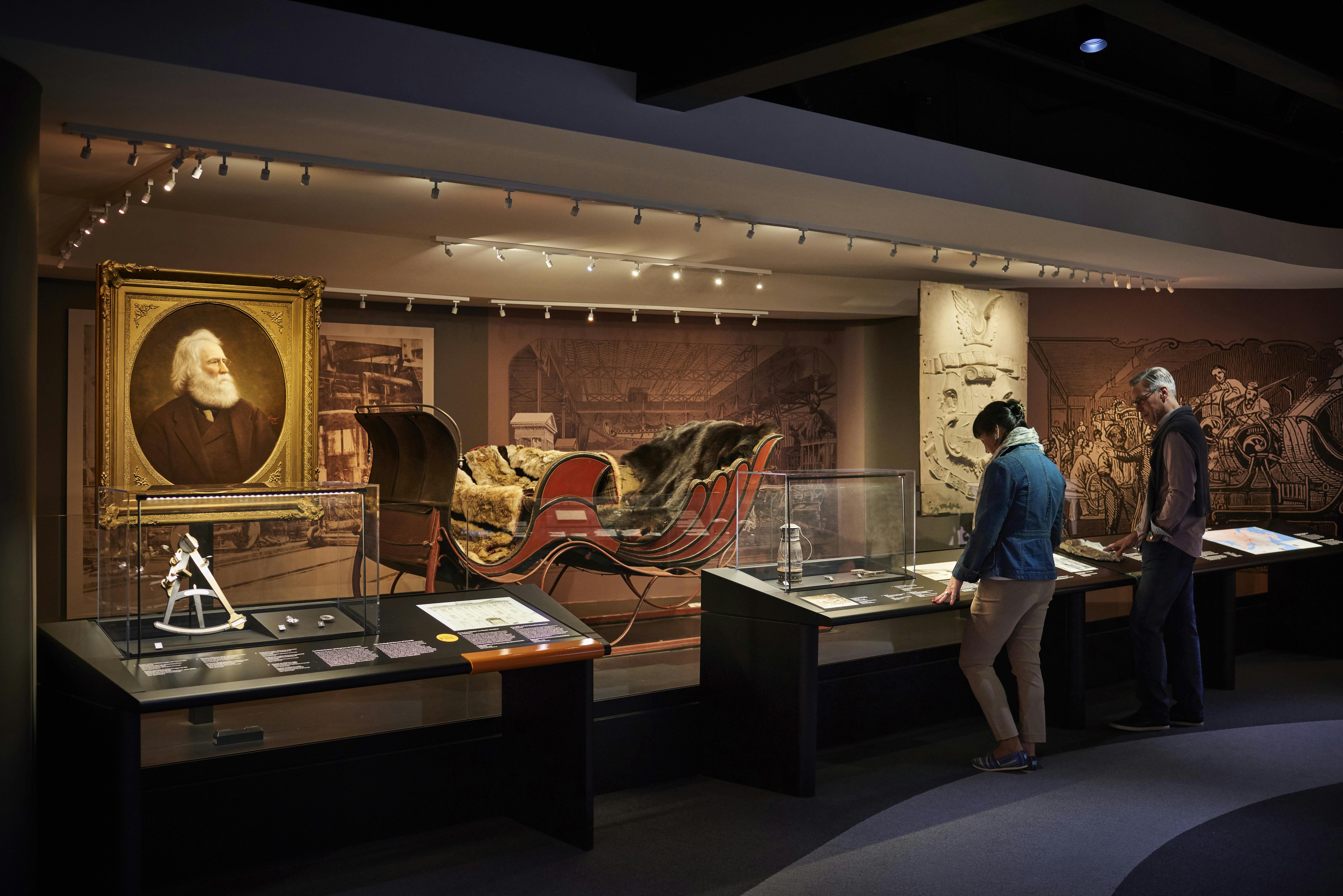 在博物馆观看展览的人，包括历史悠久的雪橇、文物和在柔和光线下拍摄的肖像。