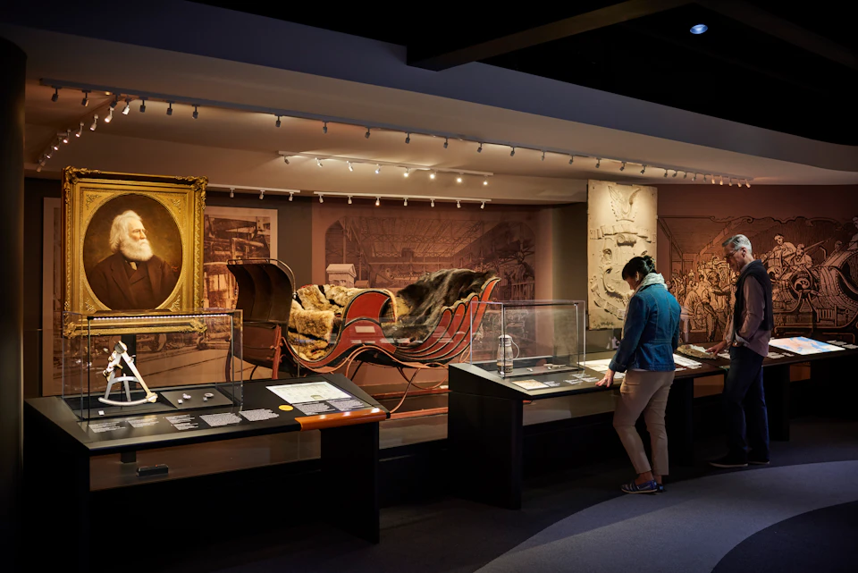 Person in museum viewing exhibits including a historic sleigh, artifacts, and framed portrait under soft lighting.