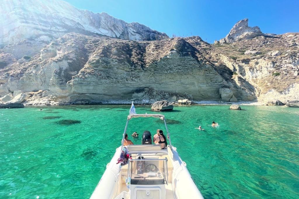 Άνθρωποι σε μια βάρκα σε καθαρά γαλαζοπράσινα νερά κοντά σε βραχώδεις βράχους. Μερικοί άνθρωποι κολυμπούν κοντά.