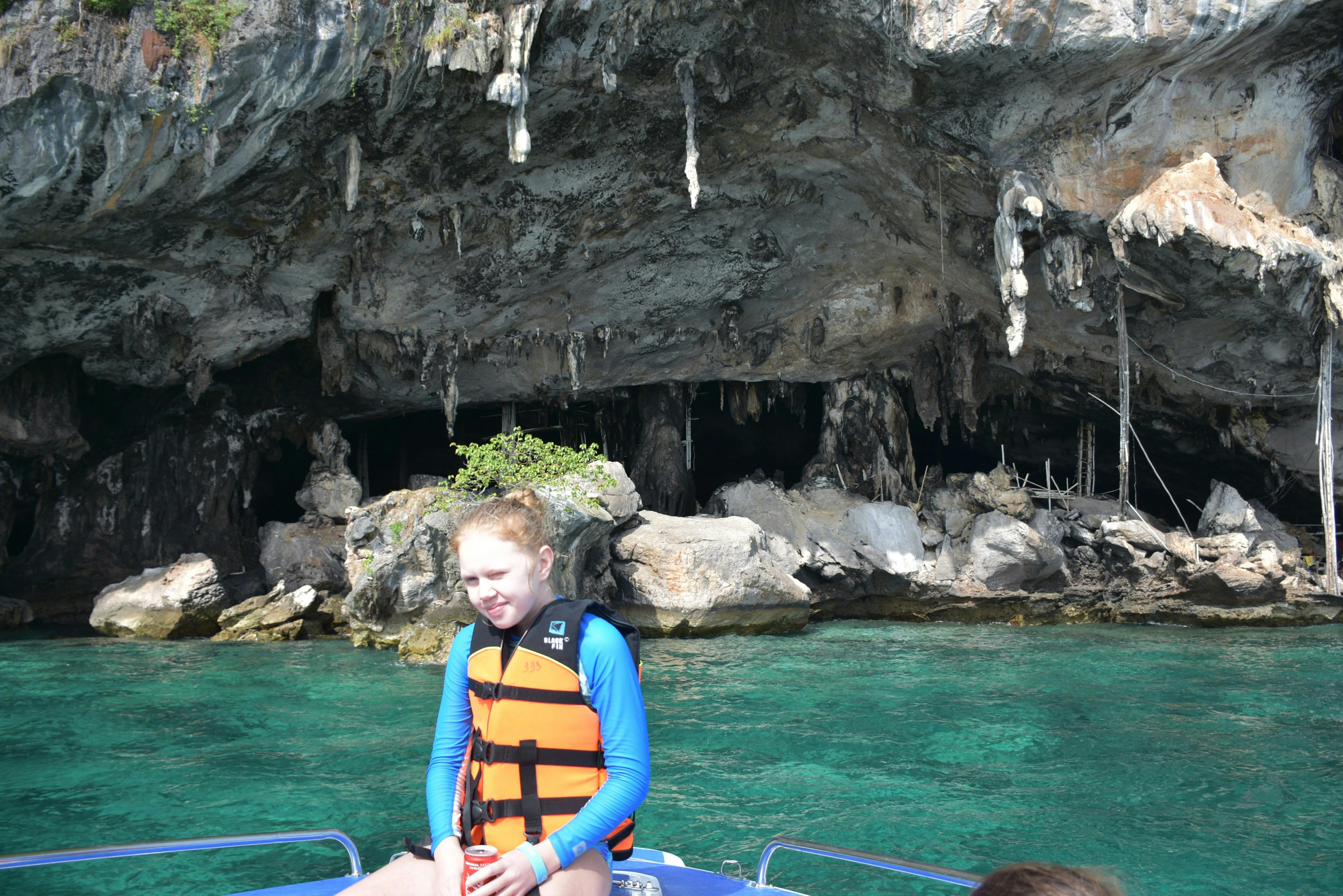 Ένα άτομο που φοράει ένα πορτοκαλί σωσίβιο κάθεται σε μια βάρκα μπροστά από βραχώδεις βράχους και καθαρά τυρκουάζ νερά.