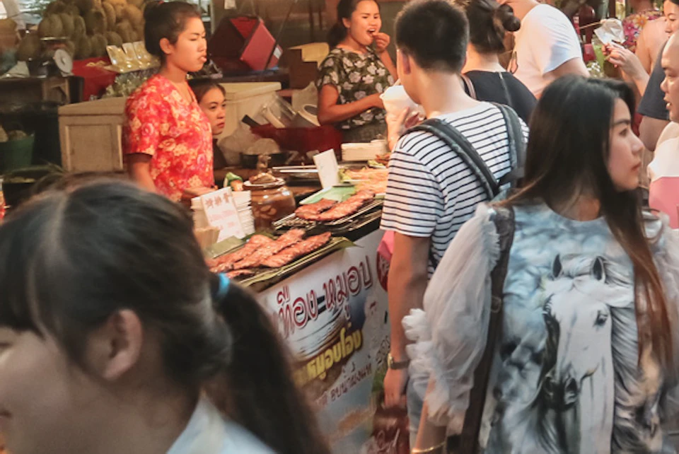 A busy market scene with people shopping at various food and goods stalls under wooden roofs.