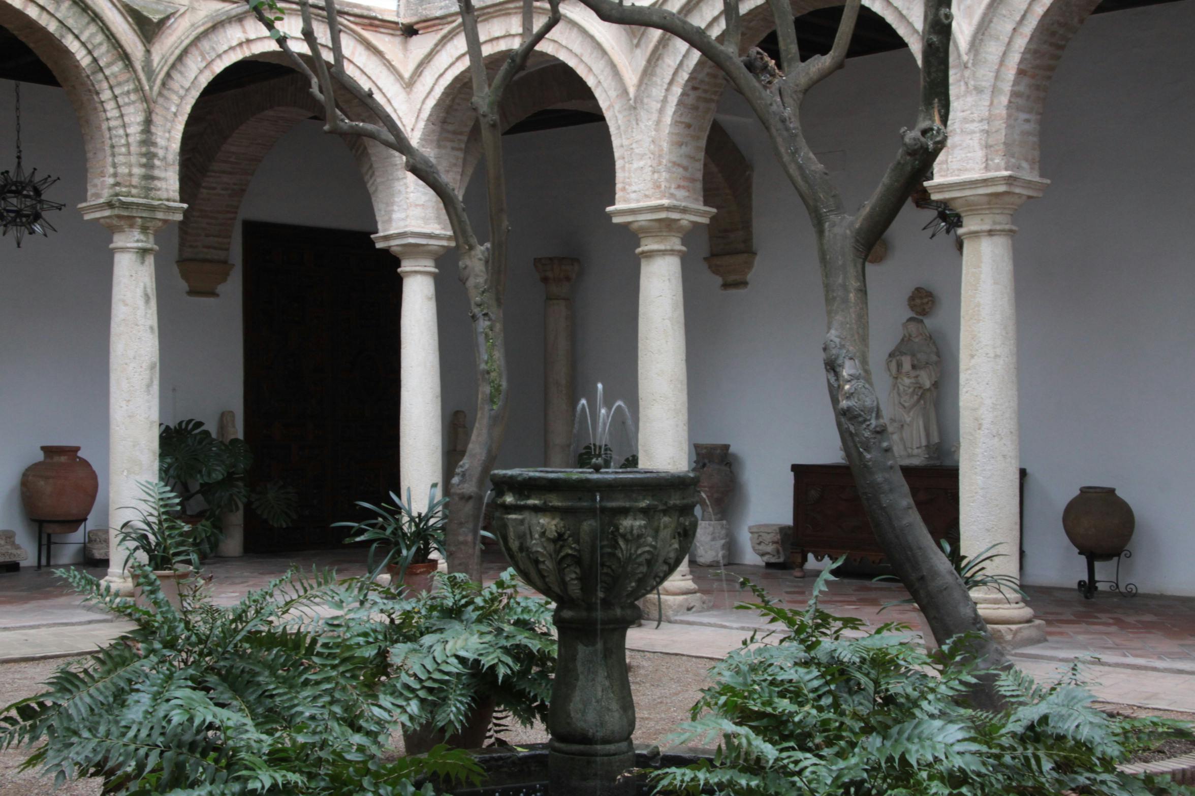 Courtyard with stone arches, a central fountain surrounded by plants, and a statue in the background.