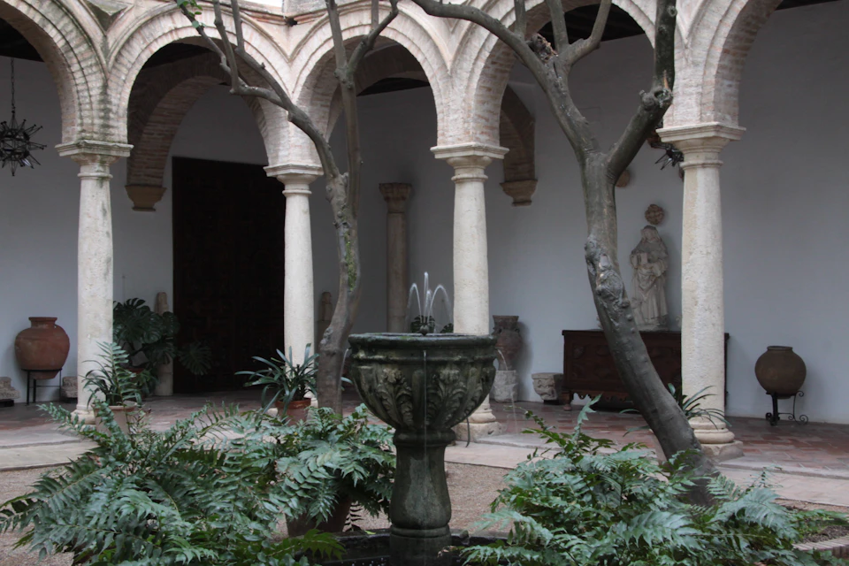 Courtyard with stone arches, a central fountain surrounded by plants, and a statue in the background.