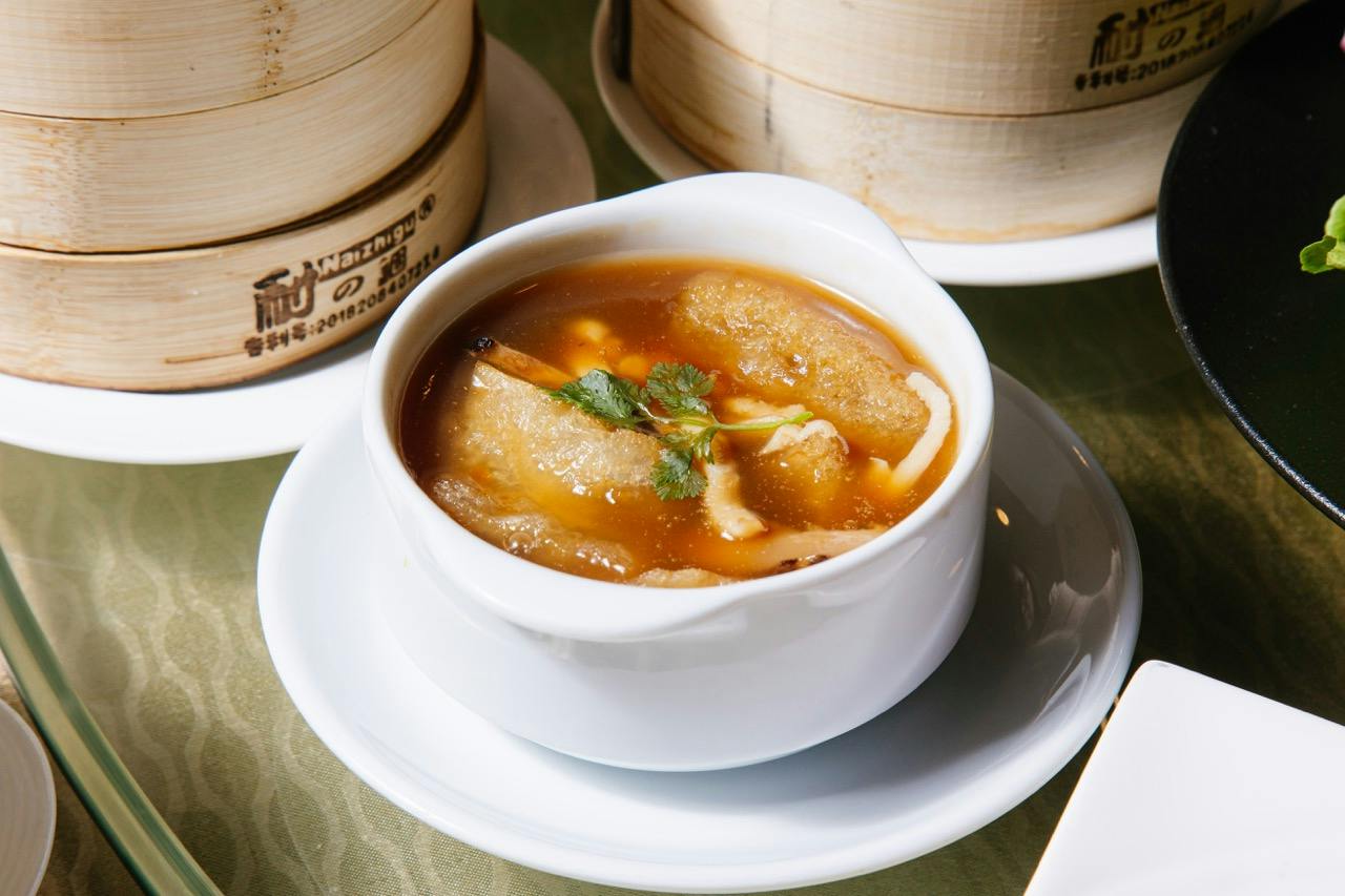 A bowl of soup with herbs and ingredients served on a saucer, with bamboo steamer baskets in the background.
