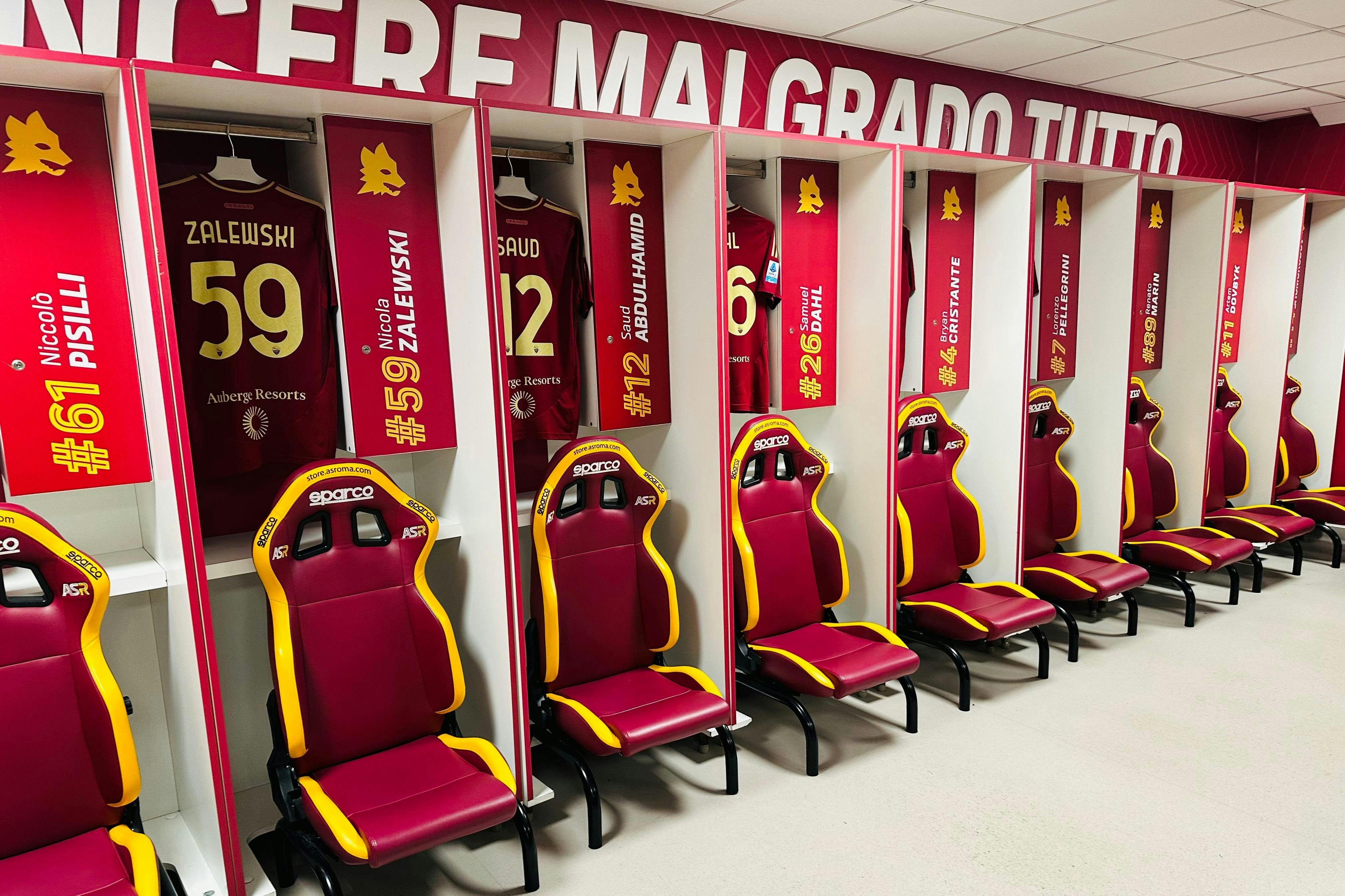 一间运动更衣室，储物柜前摆放着红色和黄色赛车风格的椅子，上面摆放着红色球衣，上面有数字和名字。