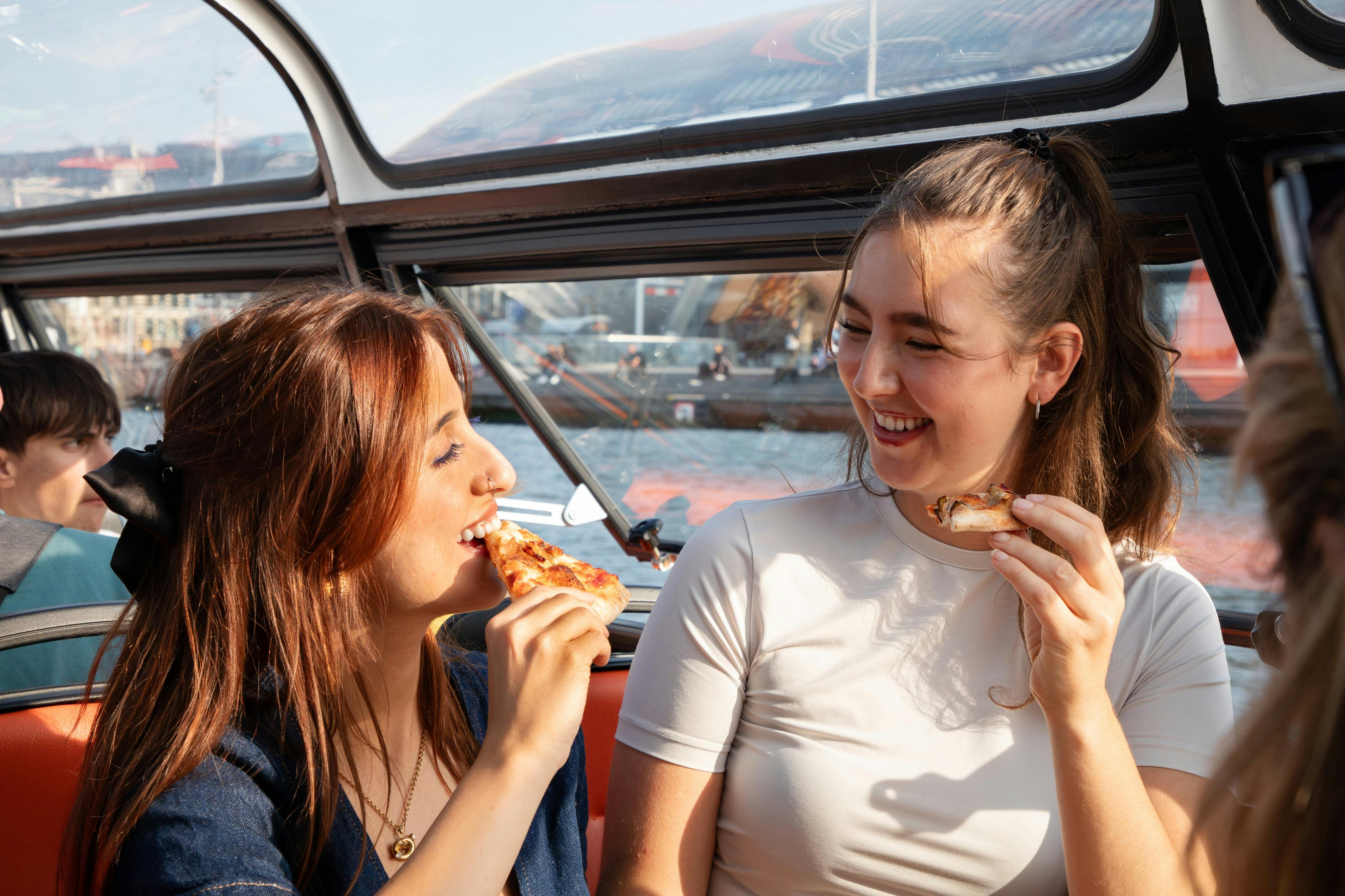세 사람이 보트 안에서 피자를 나눠 먹으며 미소를 짓고 있는데, 창문 너머로 물과 도시 풍경이 한눈에 들어옵니다.