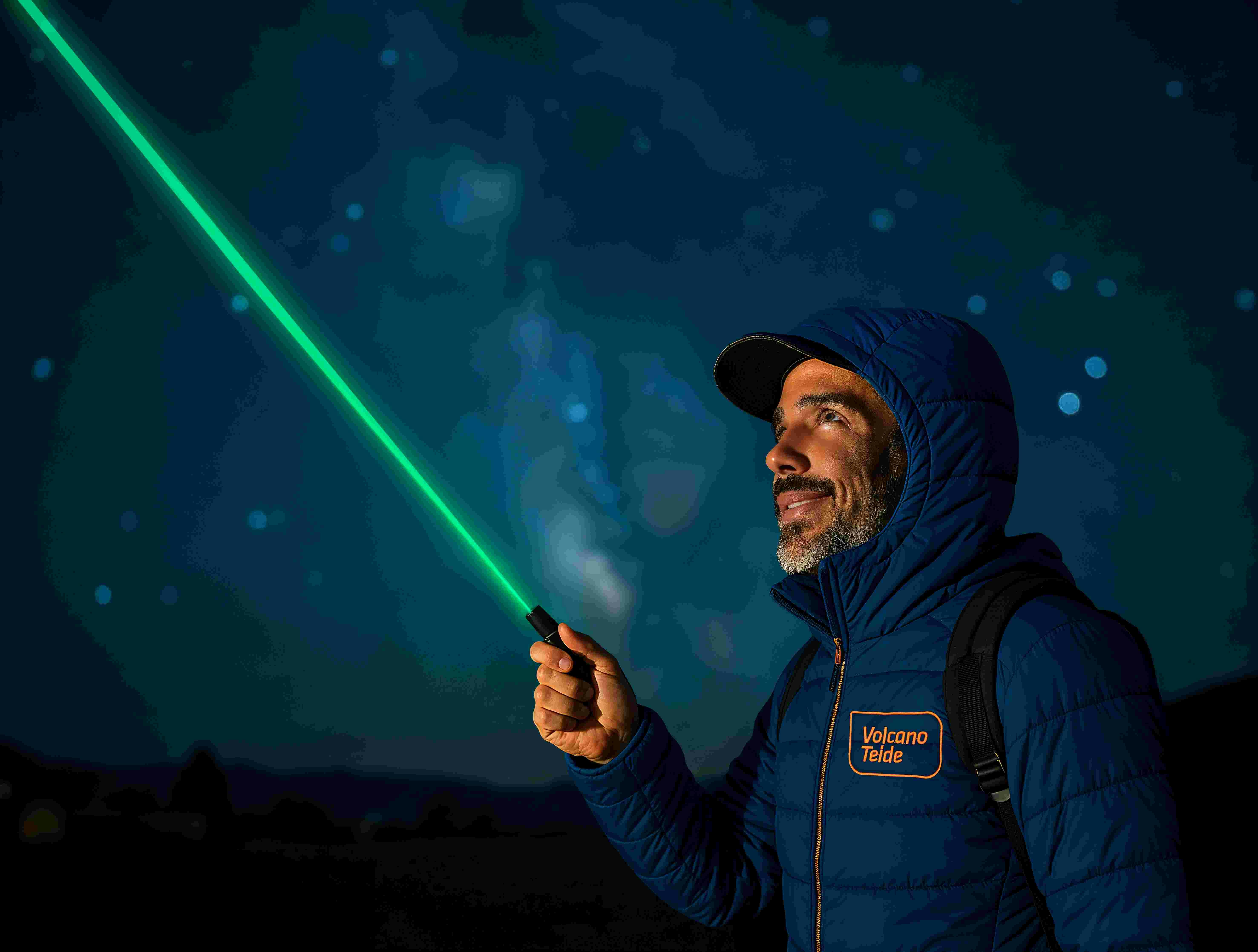 A person wearing a blue jacket points a green laser pointer at the night sky with stars visible in the background.