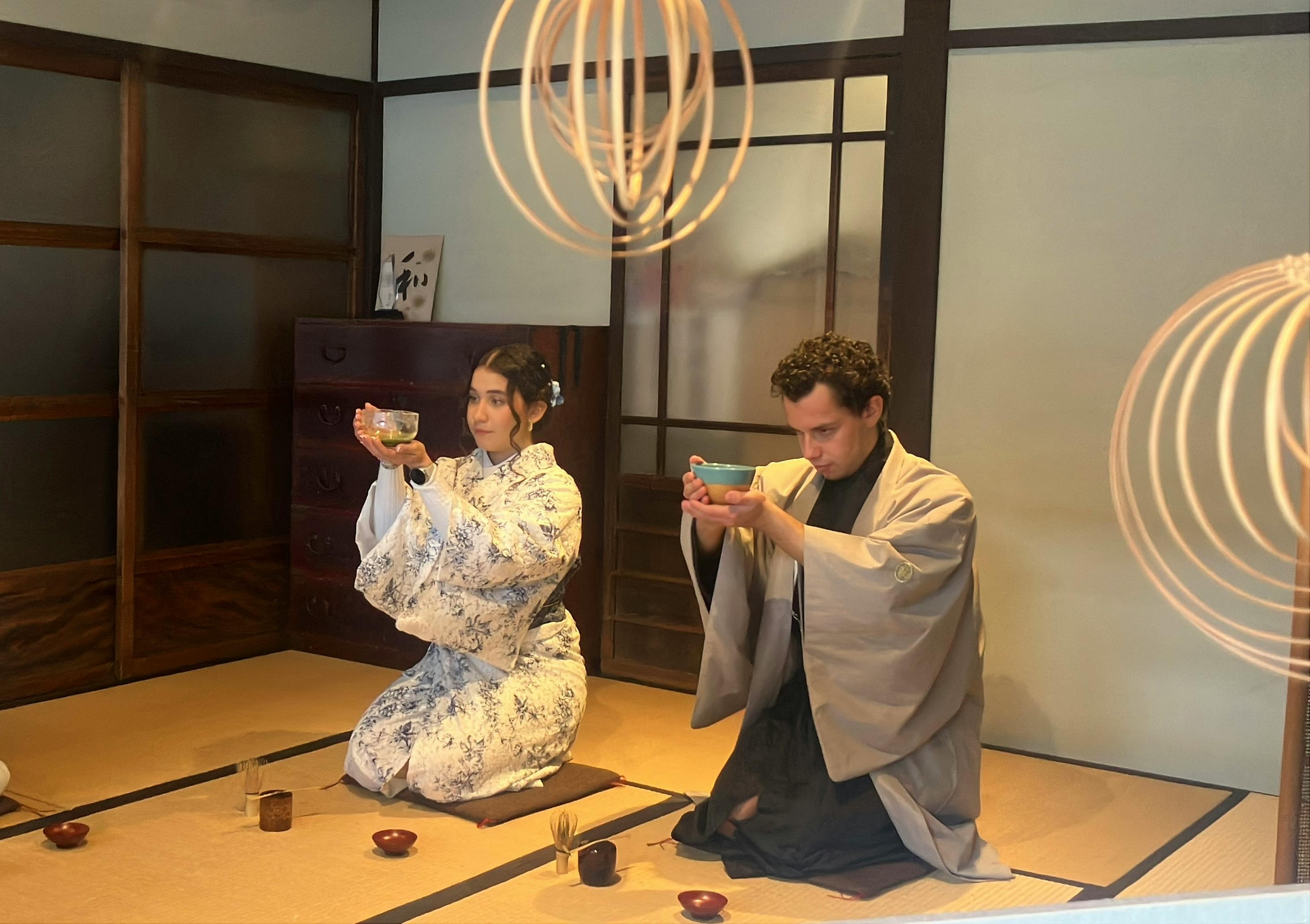 Two people in traditional Japanese attire participating in a tea ceremony in a tatami room with sliding doors.