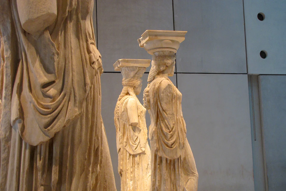 Three ancient statues of women with draped clothing and intricate hairstyles, each supporting a column on their heads.