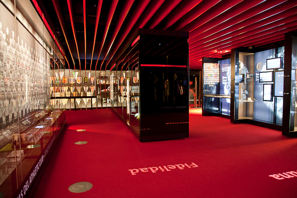 A museum exhibit with red carpet, black display cases, and various artifacts. The ceiling has red lighting and parallel beams.